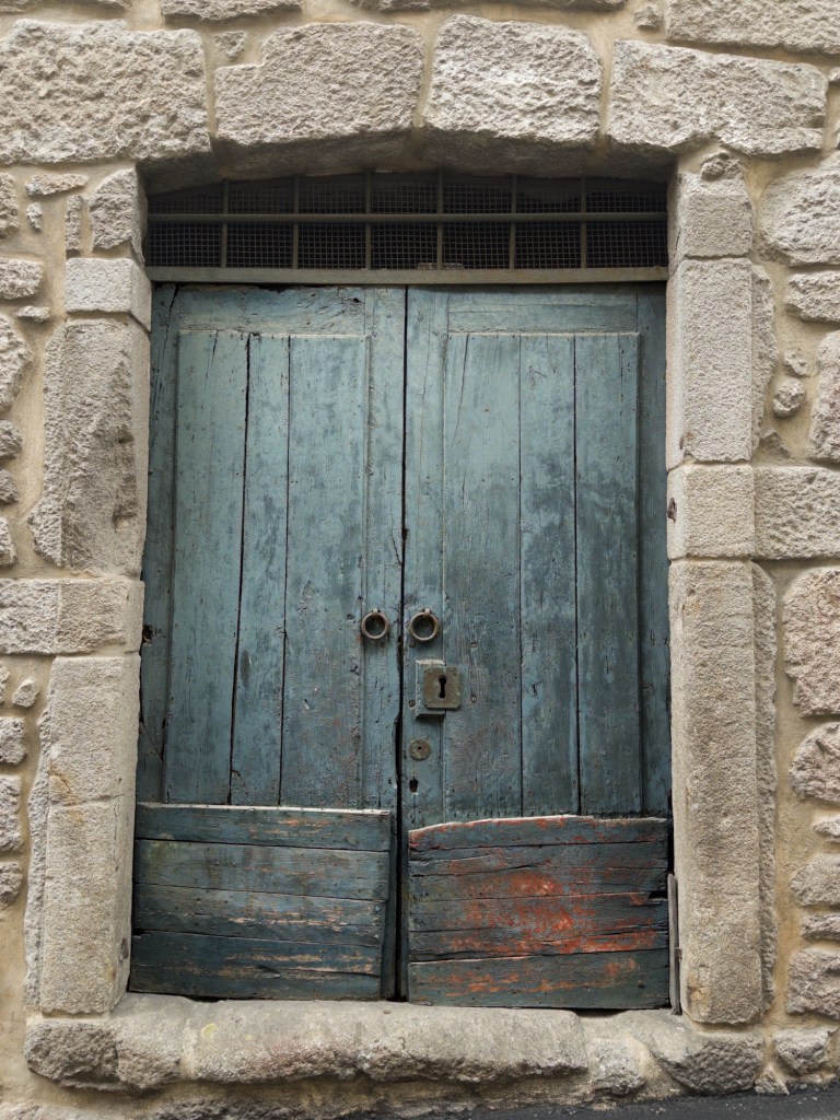 A weathered blue wooden door set in a stone wall, featuring two circular handles and a keyhole, with peeling paint and visible wear.