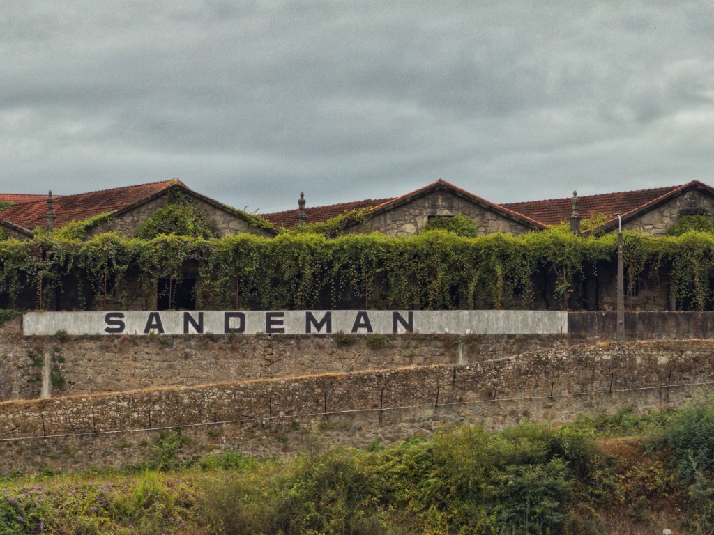 A view of a rustic building with the word 'SANDEMAN' prominently displayed on the wall, surrounded by greenery and under a cloudy sky.