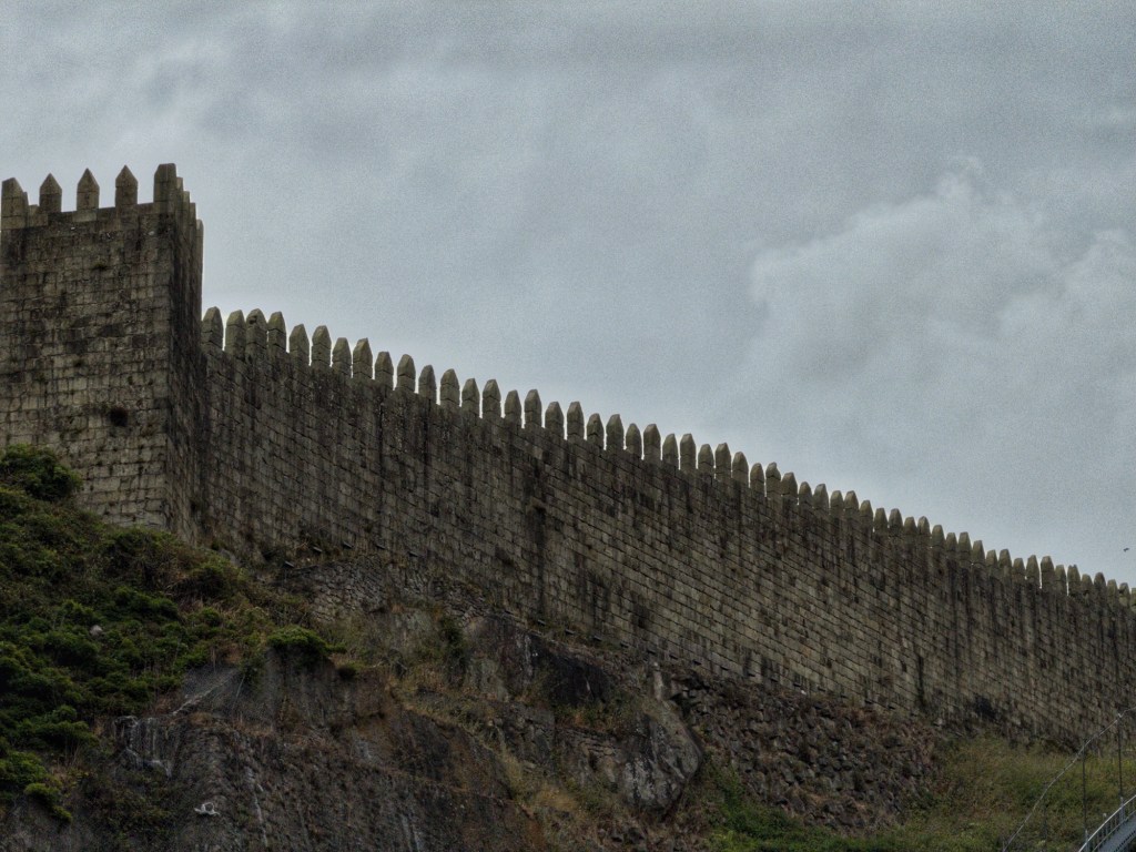 Vertical view of a medieval stone wall with crenellations, set against a cloudy sky.
