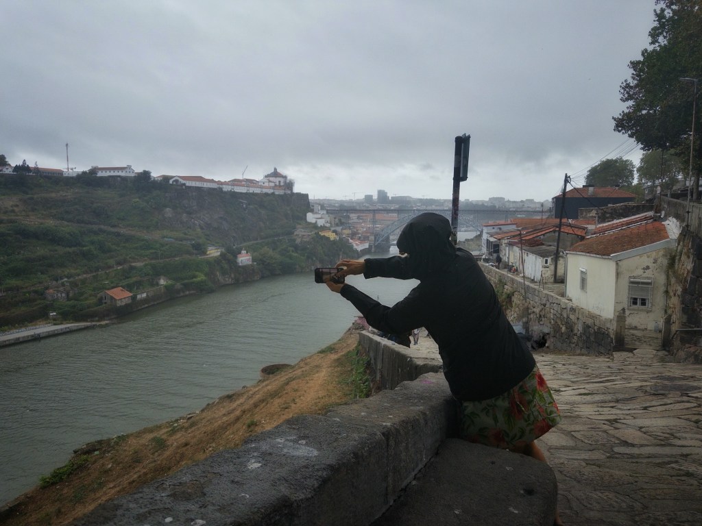 A person in a black jacket taking a photo of a river and buildings under a cloudy sky.