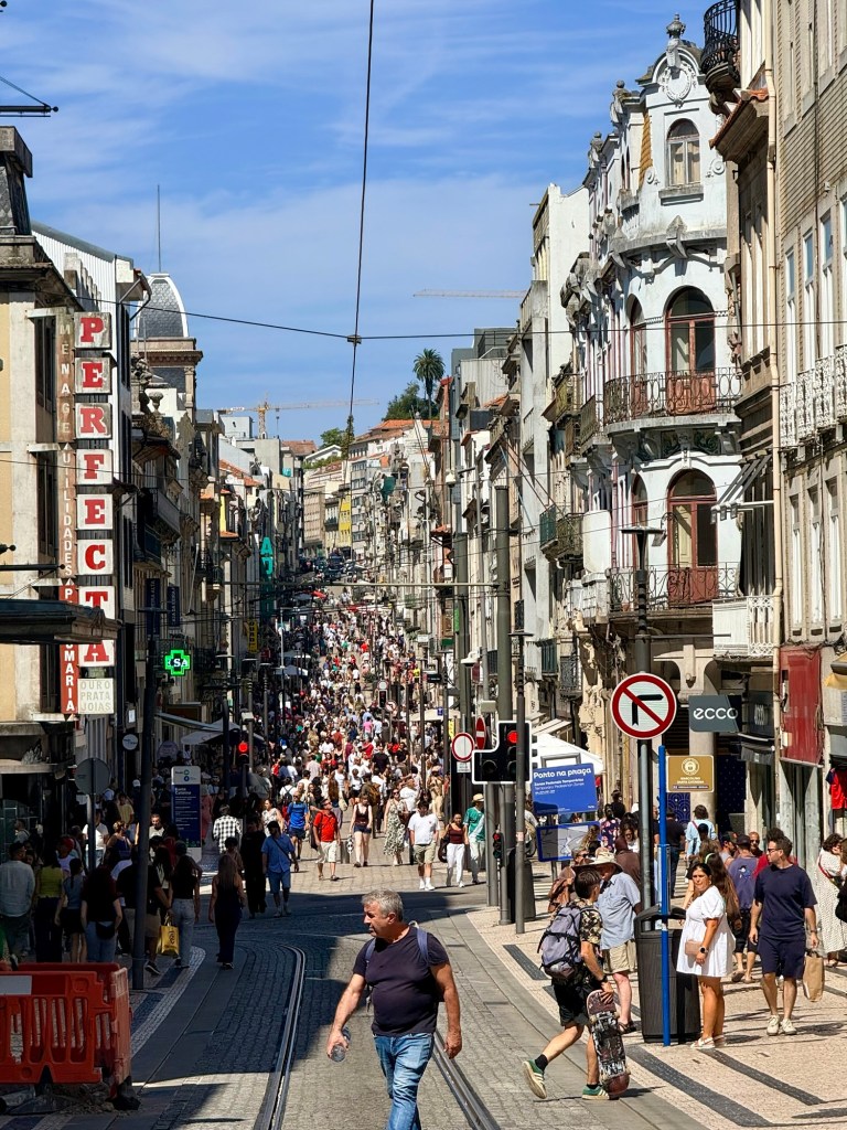 A bustling street scene in a city with numerous pedestrians, shops, and historical buildings lining both sides. The path is busy with people walking, and a clear blue sky overhead.