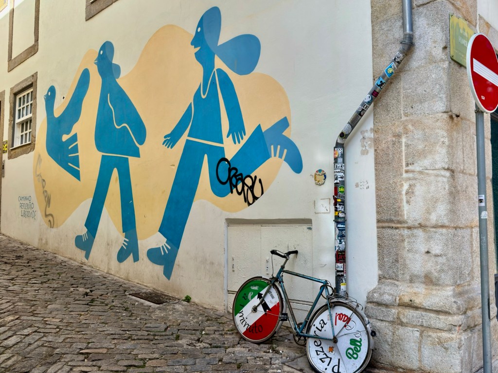 A bicycle parked against a wall with a colorful mural featuring abstract figures and a bird in blue and orange hues.
