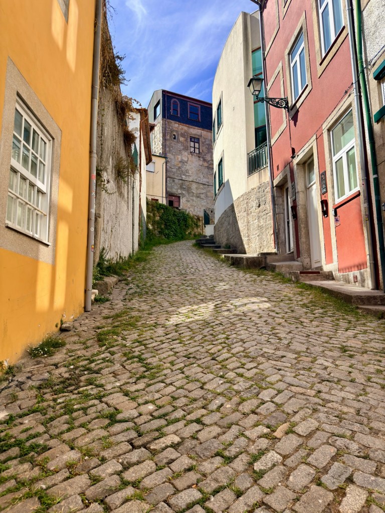 A narrow cobblestone street lined with colorful buildings, sloping uphill under a clear blue sky.