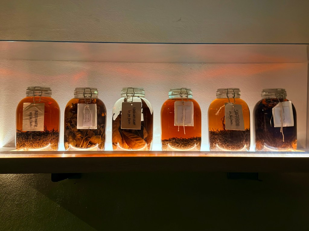 A display of six jars filled with various herbs and spices, each labeled, arranged on a shelf with a soft light illuminating them.